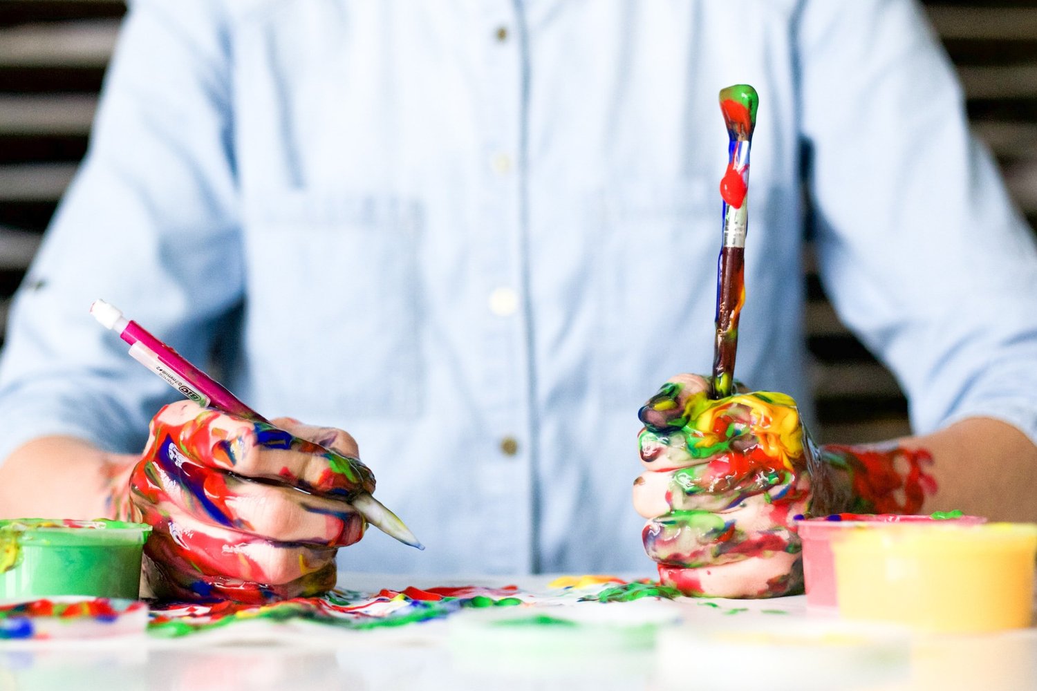 A child's hands covered in bright paint, gripping a brush and pencil